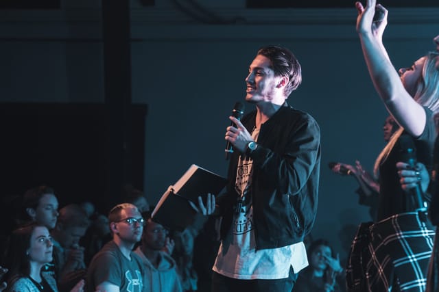 A speaker holding an open Bible and a microphone addresses a crowd of students