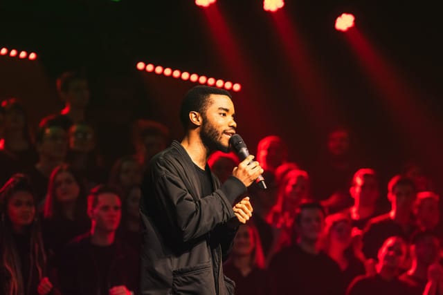 A student sings into a microphone under red stage lights with a choir behind him