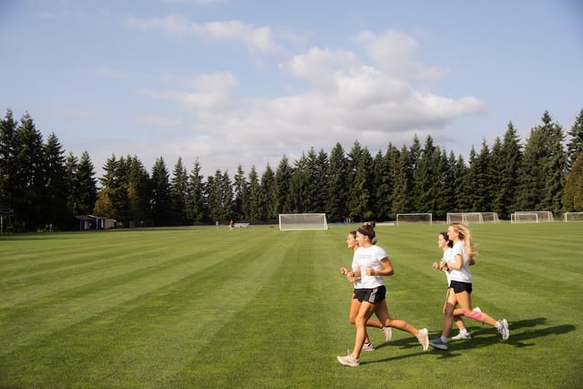 Four students jogging across the campus athletic fields, with soccer goals and evergreens behind them