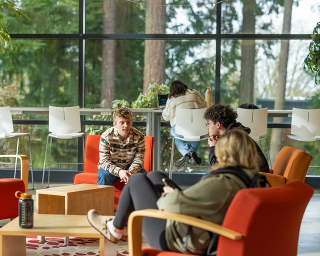 Students talking in bright orange chairs by floor-to-ceiling windows overlooking evergreen trees