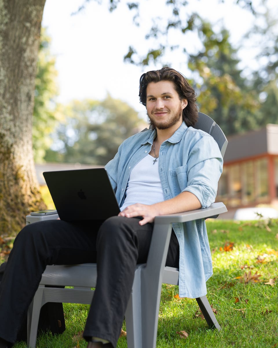 A student working on a laptop in an outdoor chair on a sunny lawn