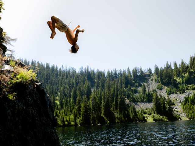 A student backflipping off a rock ledge into Snow Lake, with evergreen-covered slopes behind